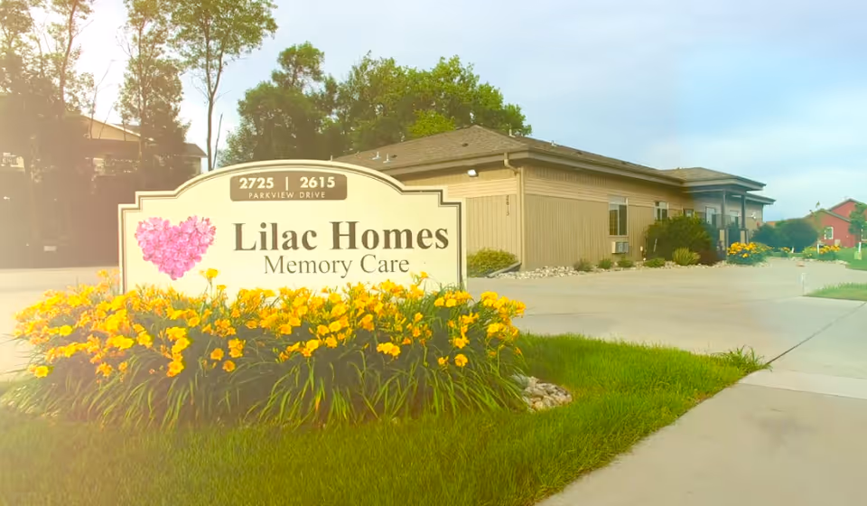 A landscaped entrance sign reading "Lilac Homes Memory Care" with yellow flowers and the facility building in the background.