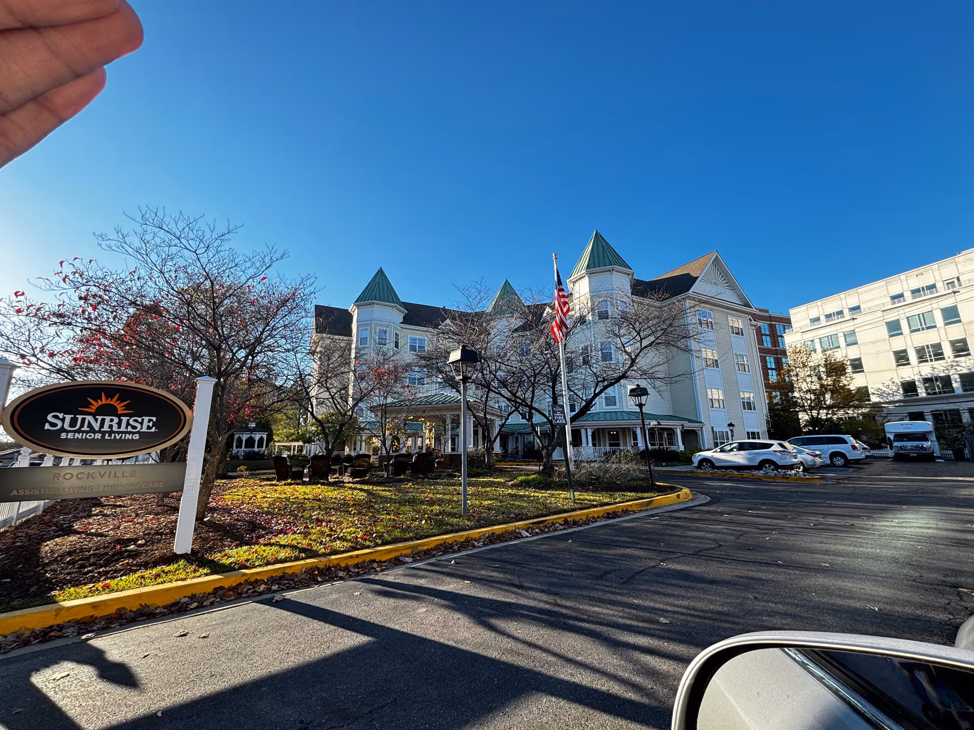 Front exterior of the Sunrise of Rockville senior living building with its entrance, sign, American flag, trees and parked cars under a clear blue sky.