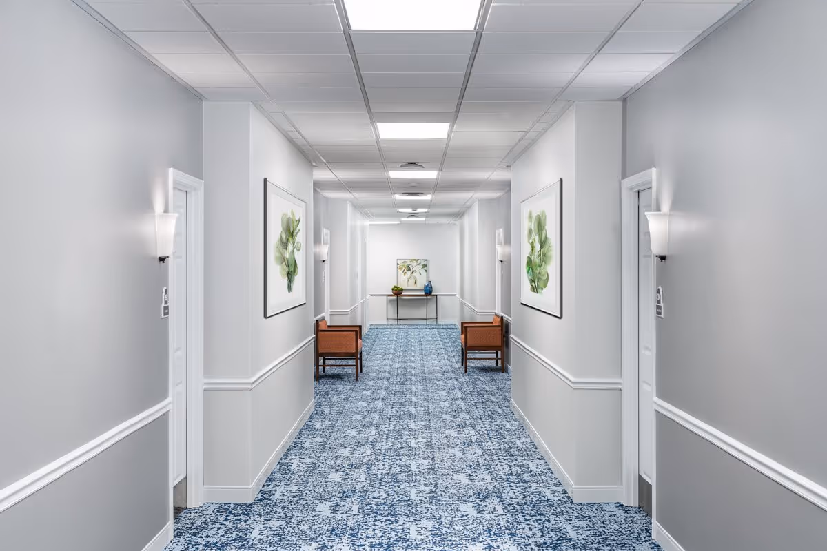 A clean, well-lit hallway in a senior living facility with light gray walls, blue patterned carpet, white doors on both sides, wall-mounted light fixtures, framed botanical artwork, and two wooden chairs with cushions near the end of the hallway beside a small table with decorative items.