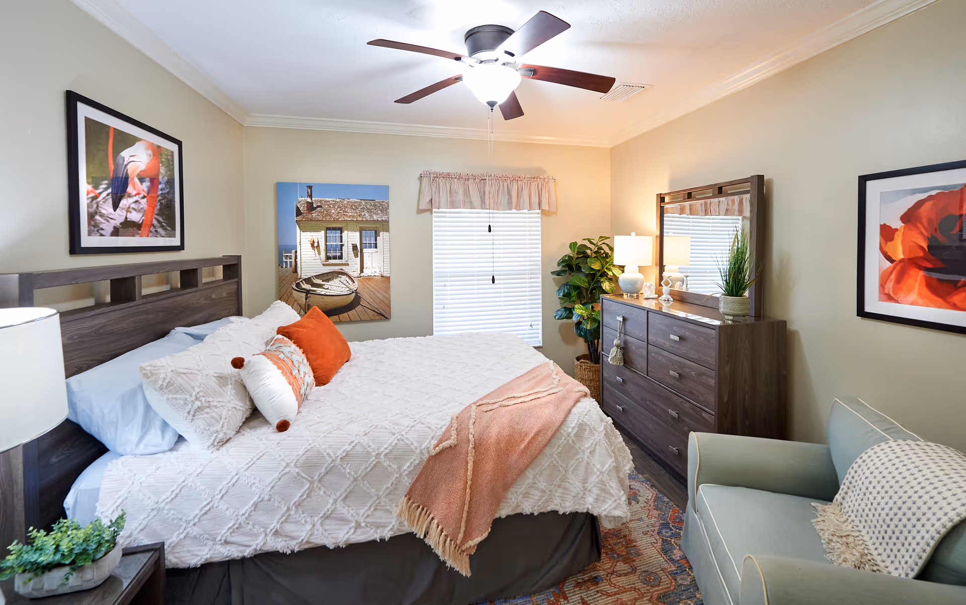 A cozy bedroom with a large bed covered in white textured bedding and orange accent pillows. The room features a ceiling fan with a light, a wooden dresser with a mirror, a green armchair with a throw blanket, and framed artwork on the walls. A window with blinds and a valance is centered on the back wall, and a potted plant is placed next to the dresser.