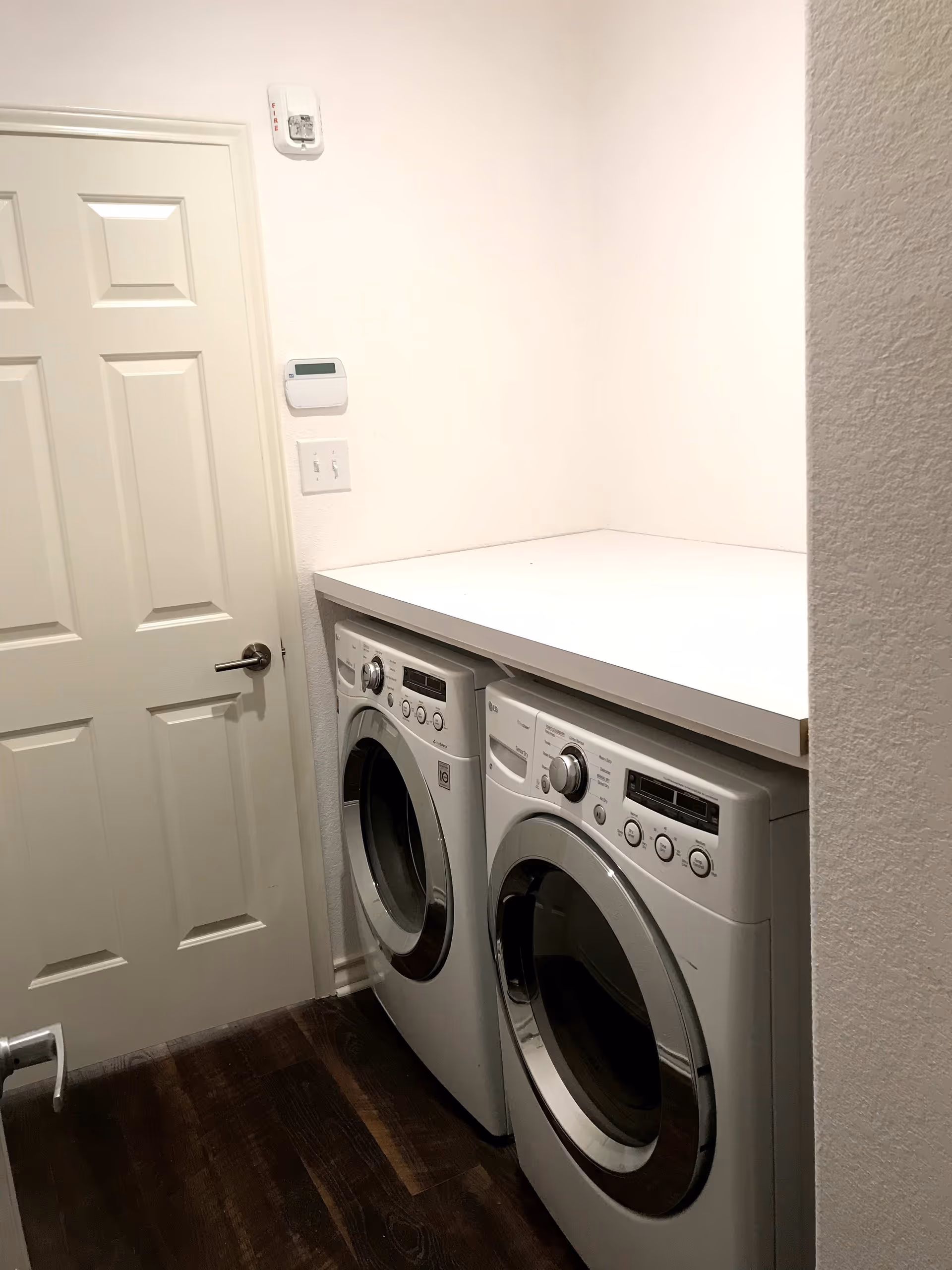 A laundry area with a front-loading washing machine and dryer side by side under a white countertop, next to a closed white door and a thermostat on the wall.