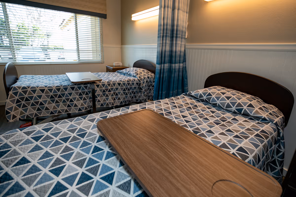 Two twin beds with geometric-patterned bedding, overbed tables, a window, and a privacy curtain in a shared senior care room.