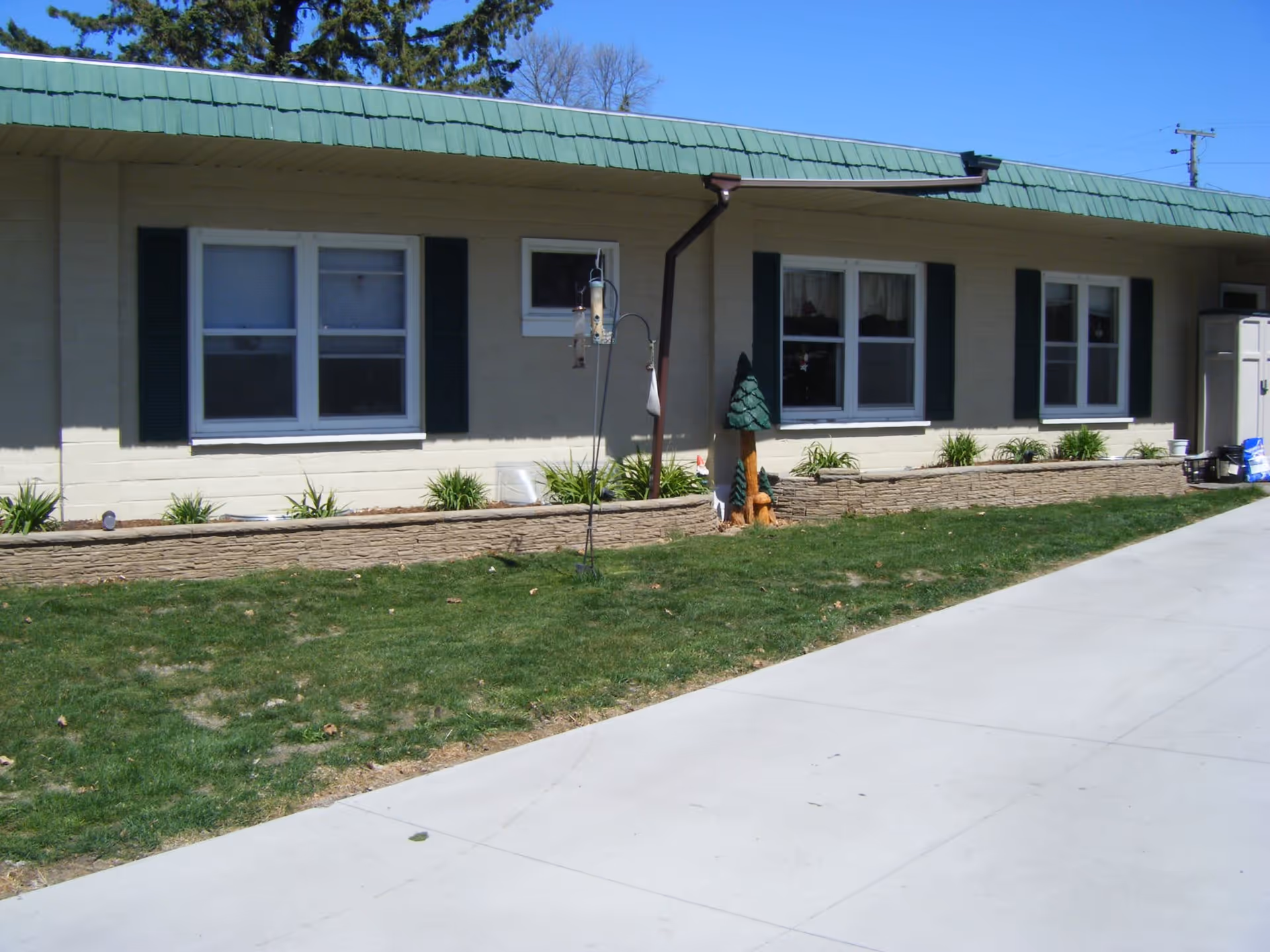 Exterior view of a single-story building with a green roof and beige walls, featuring several windows with dark green shutters. In front of the building is a small garden bed with plants and decorative wooden carvings of trees and animals. A concrete driveway runs alongside the garden and building.