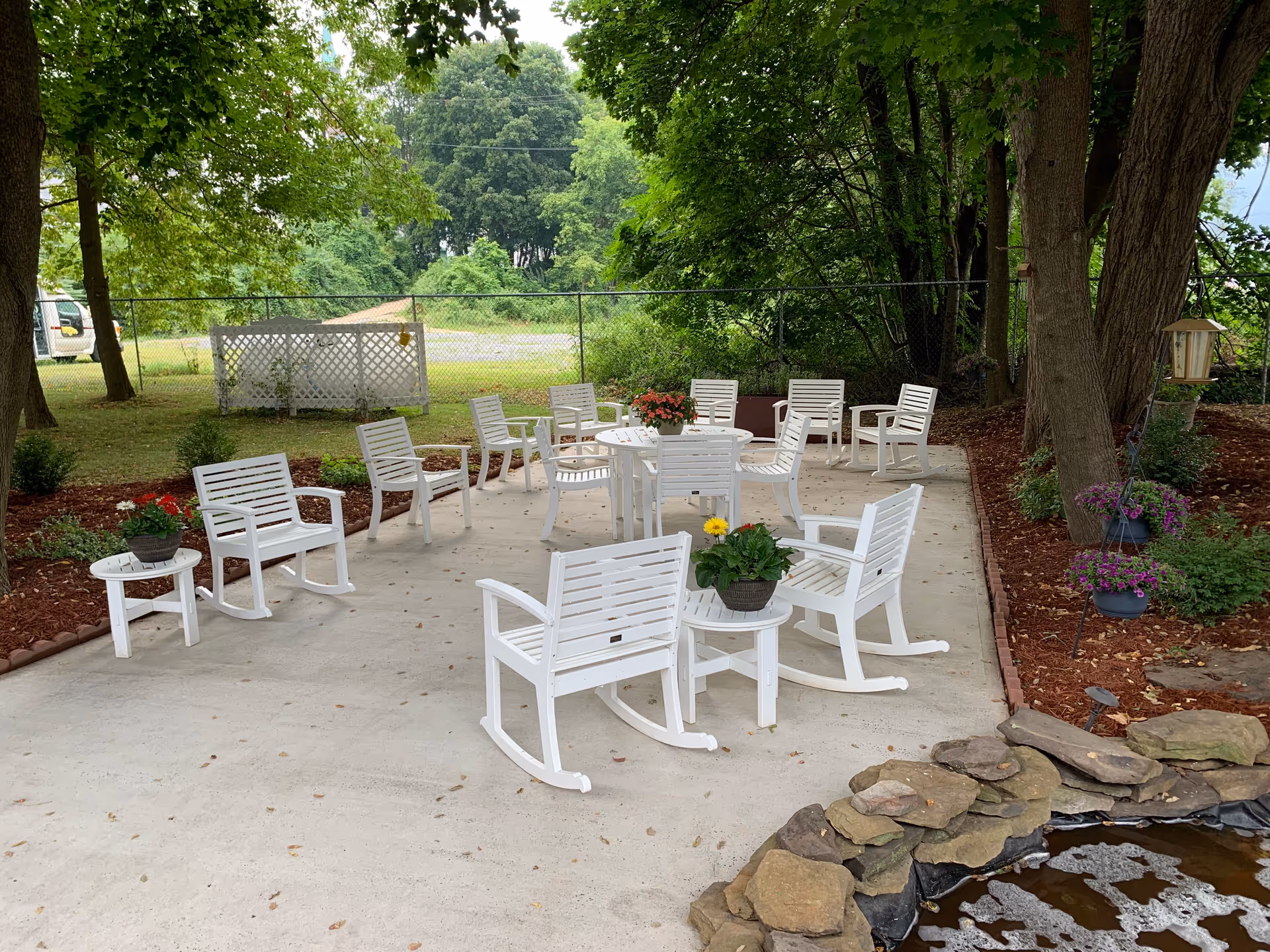 Outdoor patio with white rocking chairs and tables on a concrete pad surrounded by trees, potted plants, and a small rock-bordered pond.