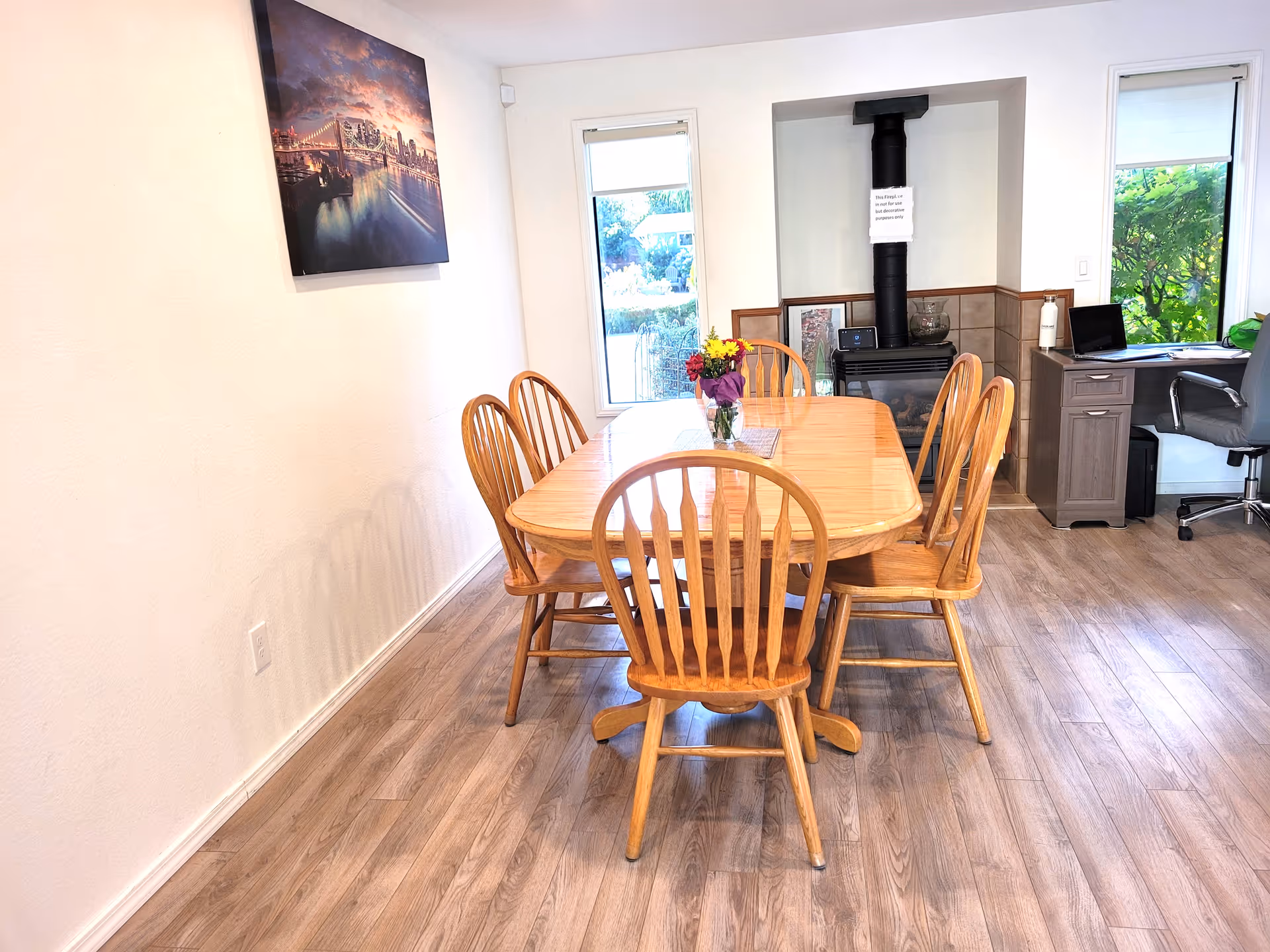 A dining room with a wooden table and six matching wooden chairs. On the table is a small vase with colorful flowers. The room has light-colored walls and wood flooring. There is a fireplace with a black chimney pipe and a sign attached to it. Two windows with white blinds let in natural light. A desk with a chair, laptop, and water bottle is situated near one window. A framed picture of a cityscape at night hangs on the wall.
