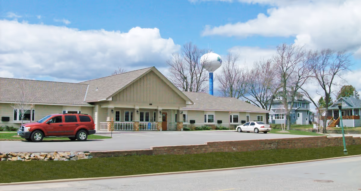 Exterior view of a single-story senior living facility building with beige siding and a gabled roof. There are two cars parked in front of the building, a red SUV and a white sedan. Leafless trees and a water tower are visible in the background under a partly cloudy blue sky.