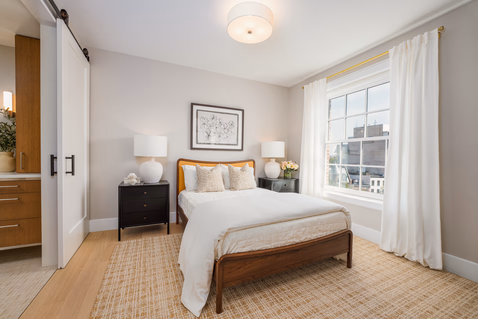A bright and neatly arranged bedroom with a wooden bed frame, white bedding, and three decorative pillows. There are two black nightstands on either side of the bed, each with a white lamp. A framed abstract artwork hangs above the bed. The room has a large window with white curtains letting in natural light, and a beige patterned rug covers the wooden floor. A sliding door on the left side leads to an adjacent room with wooden cabinetry.