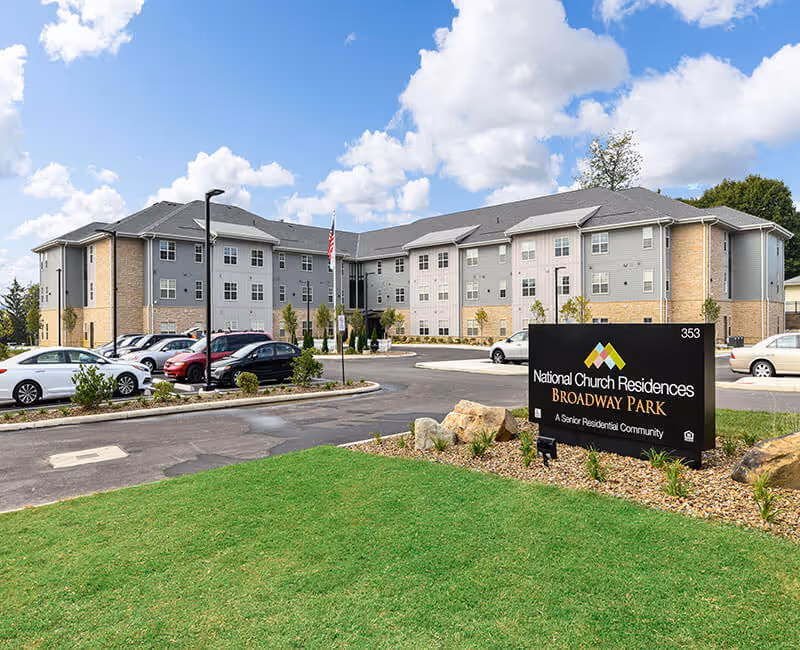 Front exterior of a three-story senior living building with parked cars, landscaping, and a prominent sign for National Church Residences Broadway Park.