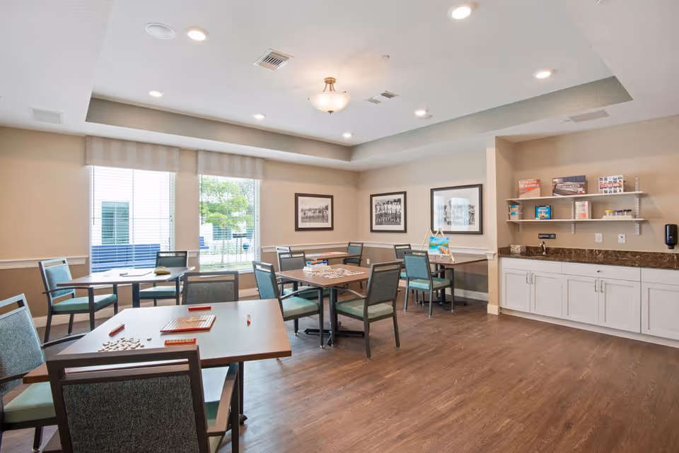 Bright activity/dining room with several tables and chairs, windows, framed pictures, and a countertop with cabinets and shelves.