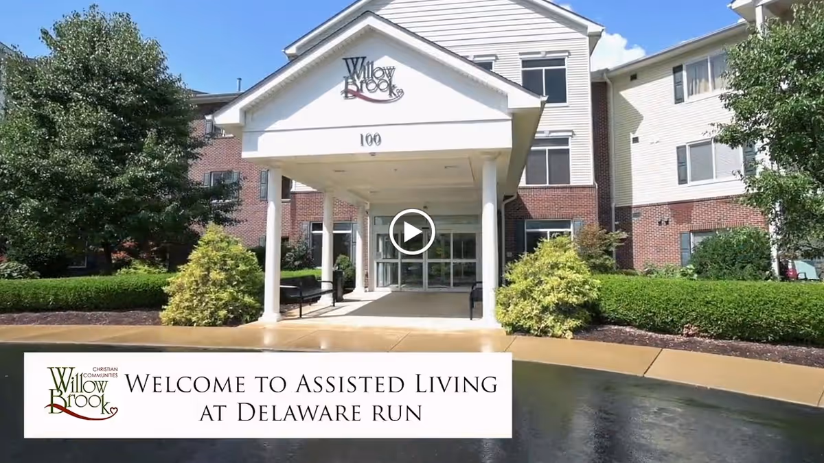 Front exterior view of Willow Brook at Delaware Run assisted living facility with a covered entrance, surrounded by greenery and bushes under a clear blue sky.