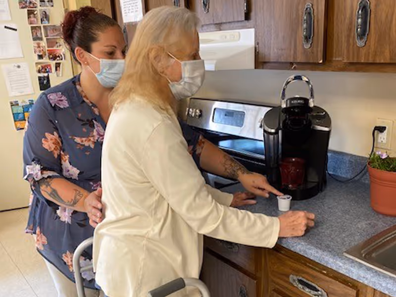 An elderly woman wearing a mask is using a coffee machine in a kitchen while being assisted by a caregiver who is also wearing a mask. The kitchen has wooden cabinets, a countertop with a coffee maker, and a small potted plant.