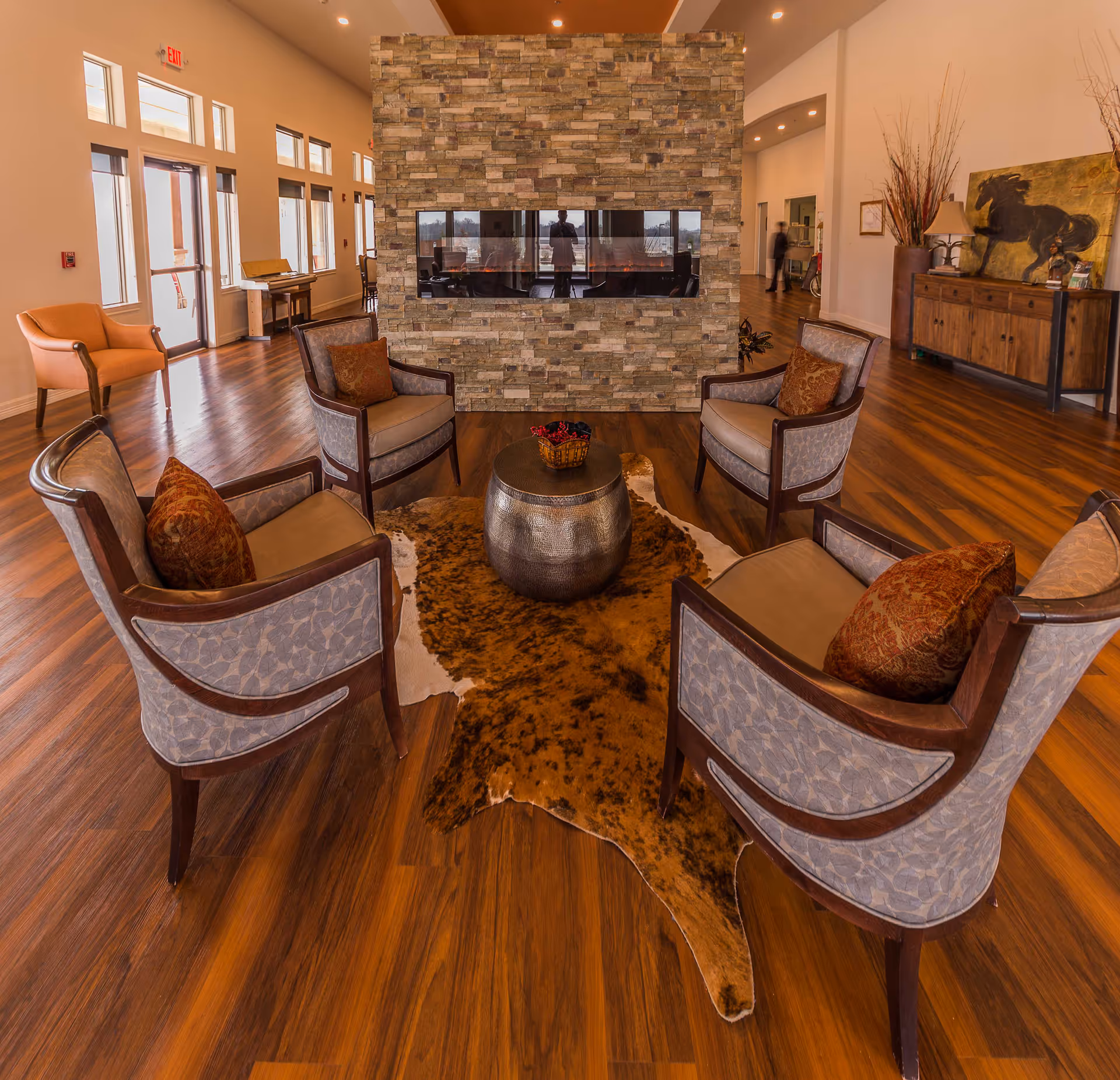 A cozy seating area in a senior living facility with four upholstered armchairs arranged around a small round metallic table on a brown animal hide rug. Behind the seating area is a stone fireplace with a glass front. The room has wooden flooring, large windows, and warm lighting. In the background, there is a wooden sideboard with a lamp and a painting of a horse.