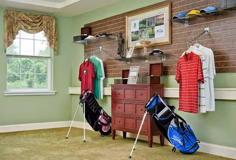 Interior room displaying golf equipment and apparel with two golf bags, hanging shirts, a wooden chest of drawers and shelving beneath a window.