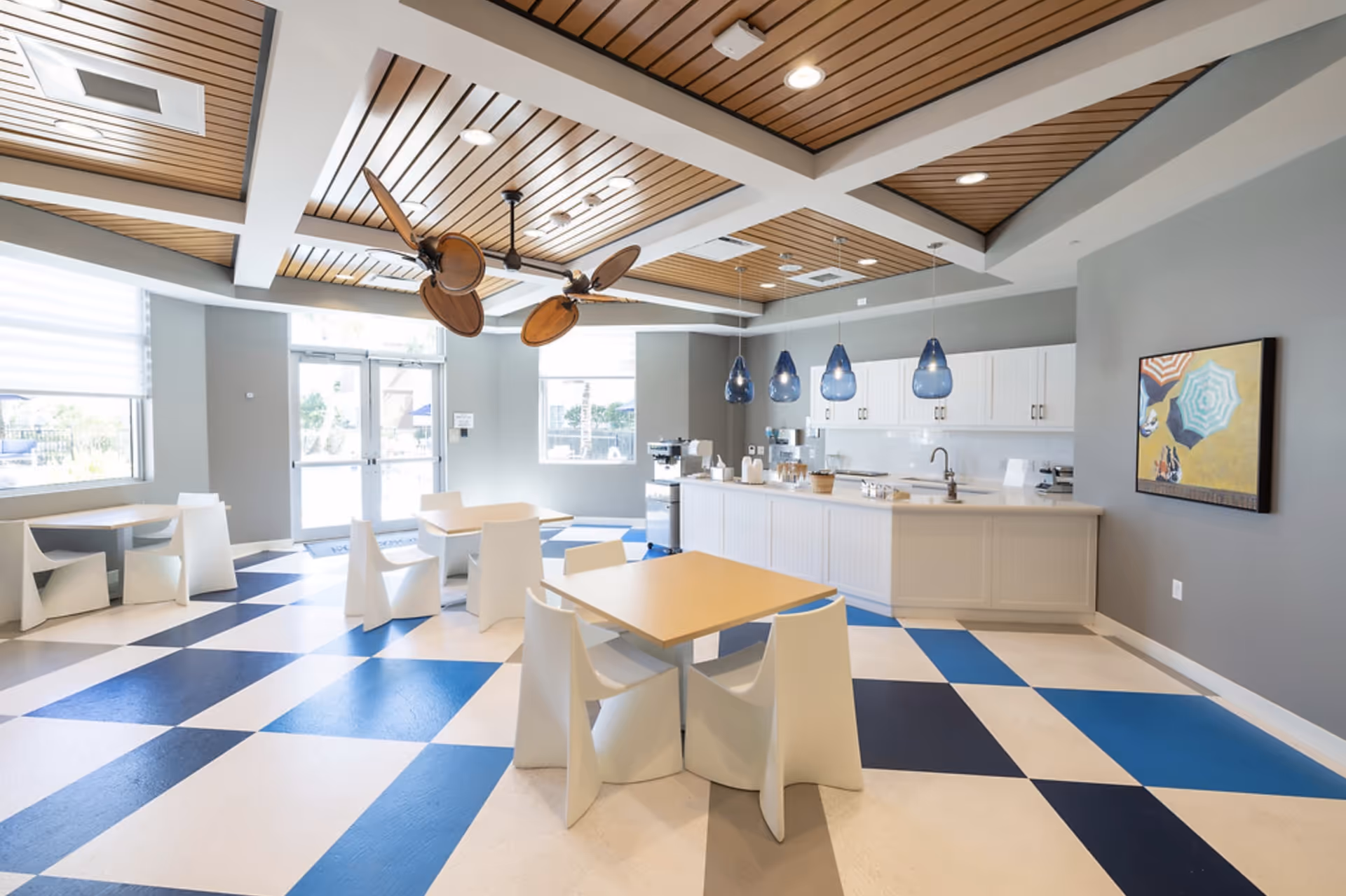 A bright and modern dining area with a checkered blue and white floor, light wood tables, and white chairs. The ceiling features wooden panels with large ceiling fans and recessed lighting. There is a kitchen counter with white cabinets and blue pendant lights hanging above it. A colorful painting is mounted on the gray wall to the right, and large windows and glass doors allow natural light to fill the space.