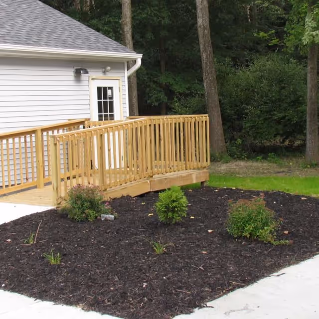 Wooden accessibility ramp leading to a side door of a light-colored building with a mulched planting bed and trees behind it.