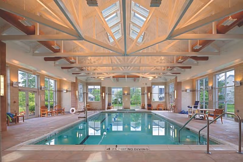 Indoor community swimming pool in a high-ceilinged room with large windows and seating around the deck.