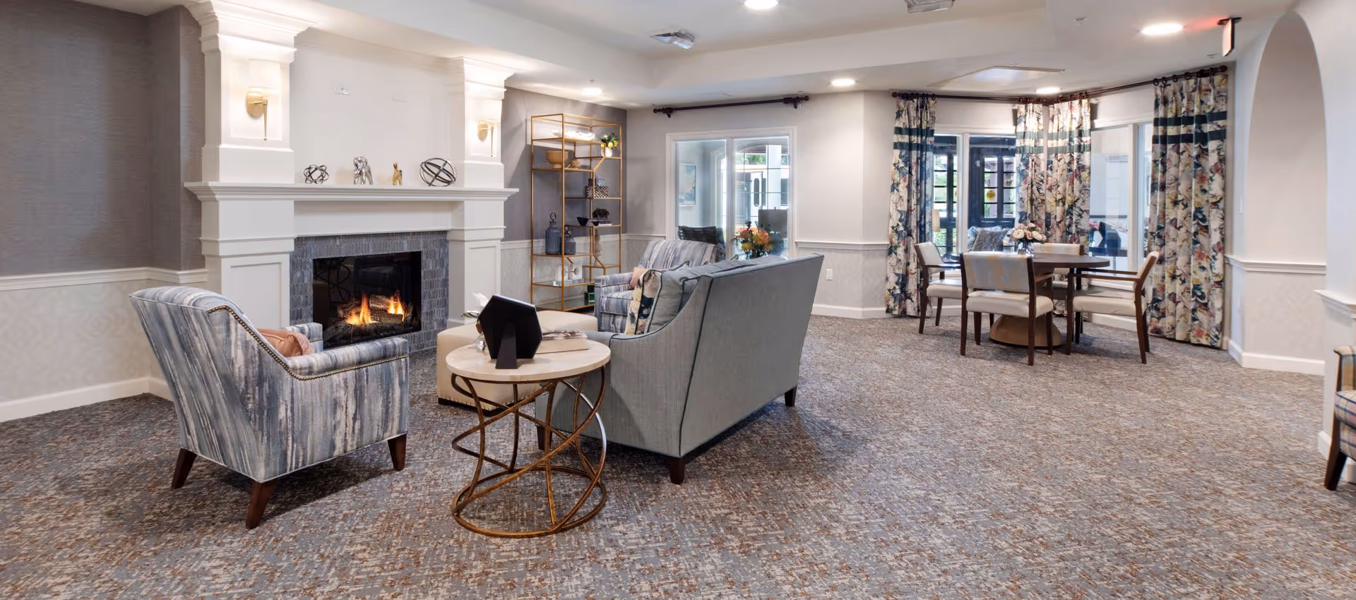Spacious communal living room with a lit fireplace, armchairs and sofa around a coffee table and a dining table by the windows.