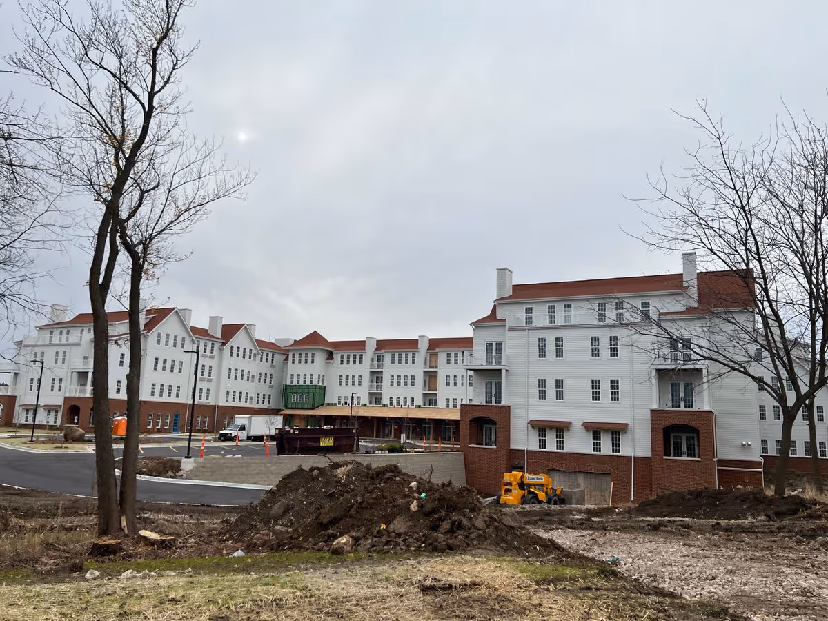 Exterior view of a large multi-story white building with red roofs under construction, with dirt piles, construction equipment, and bare trees in the foreground.