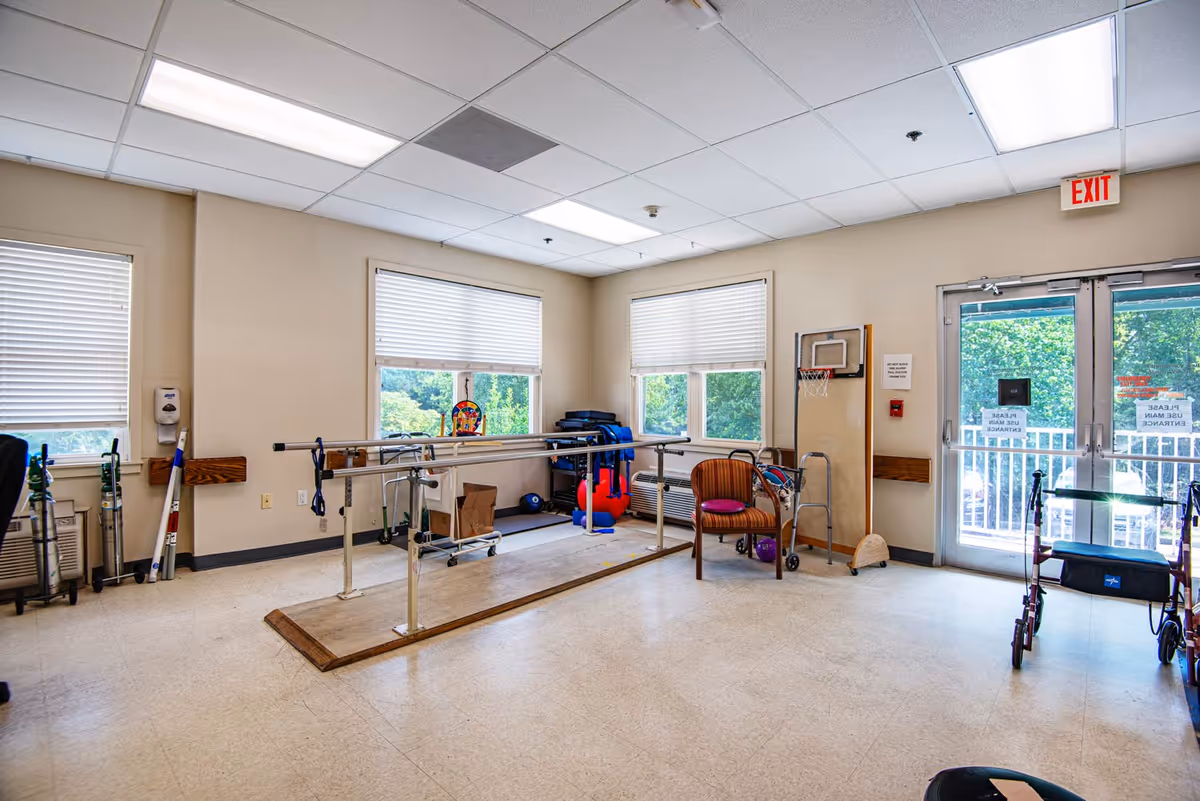 A rehabilitation room with parallel bars for walking exercises, a chair, walkers, exercise balls, and a small basketball hoop mounted on a wooden board. The room has large windows with blinds, beige walls, and a double glass door leading outside with signs on it. The floor is light-colored linoleum and the ceiling has fluorescent lights.