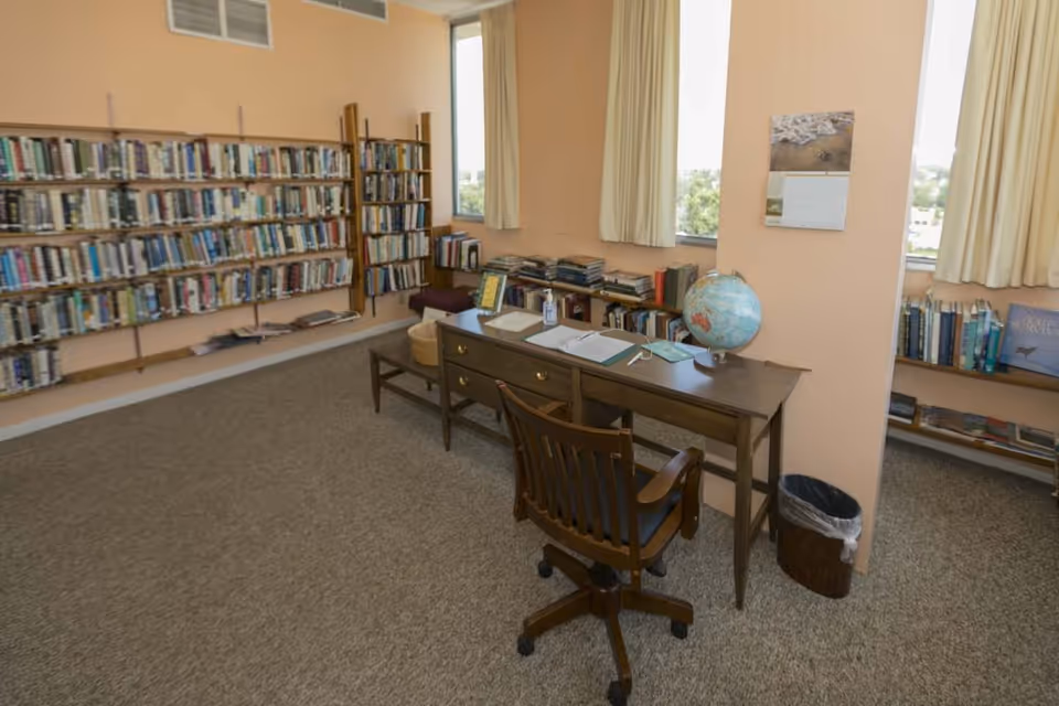 A quiet library-like room with bookshelves along the walls, a desk topped with a globe and papers, and a rolling wooden chair by curtained windows.