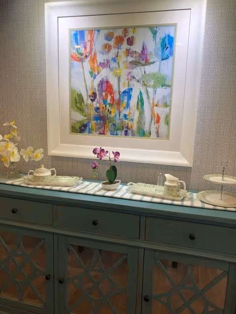 A decorative sideboard with a blue-green finish and glass-paneled doors. On top of the sideboard is a striped cloth with a small potted orchid, a teapot, a creamer, and a tiered serving tray. Above the sideboard hangs a colorful abstract painting in a white frame. The wall behind has a subtle patterned wallpaper.
