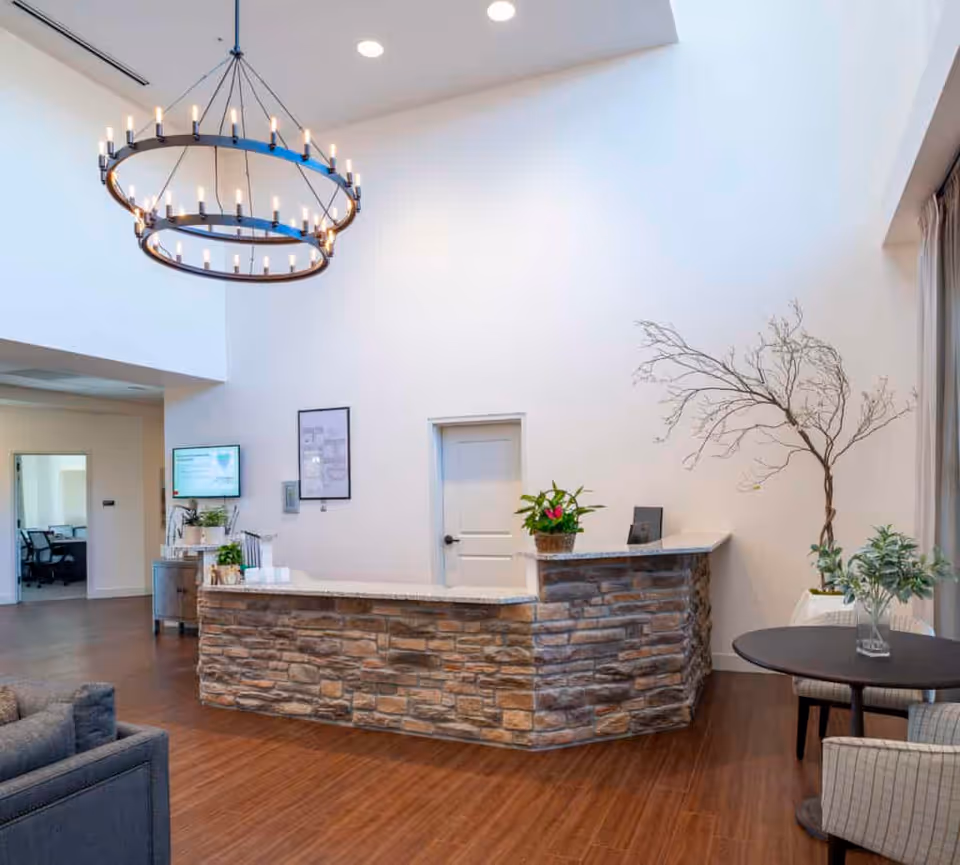 Reception area with a stone-faced front desk, a large circular chandelier with multiple lights hanging from a high ceiling, a small round table with two chairs, and decorative plants including a bare branch arrangement. The space has wooden flooring and white walls.