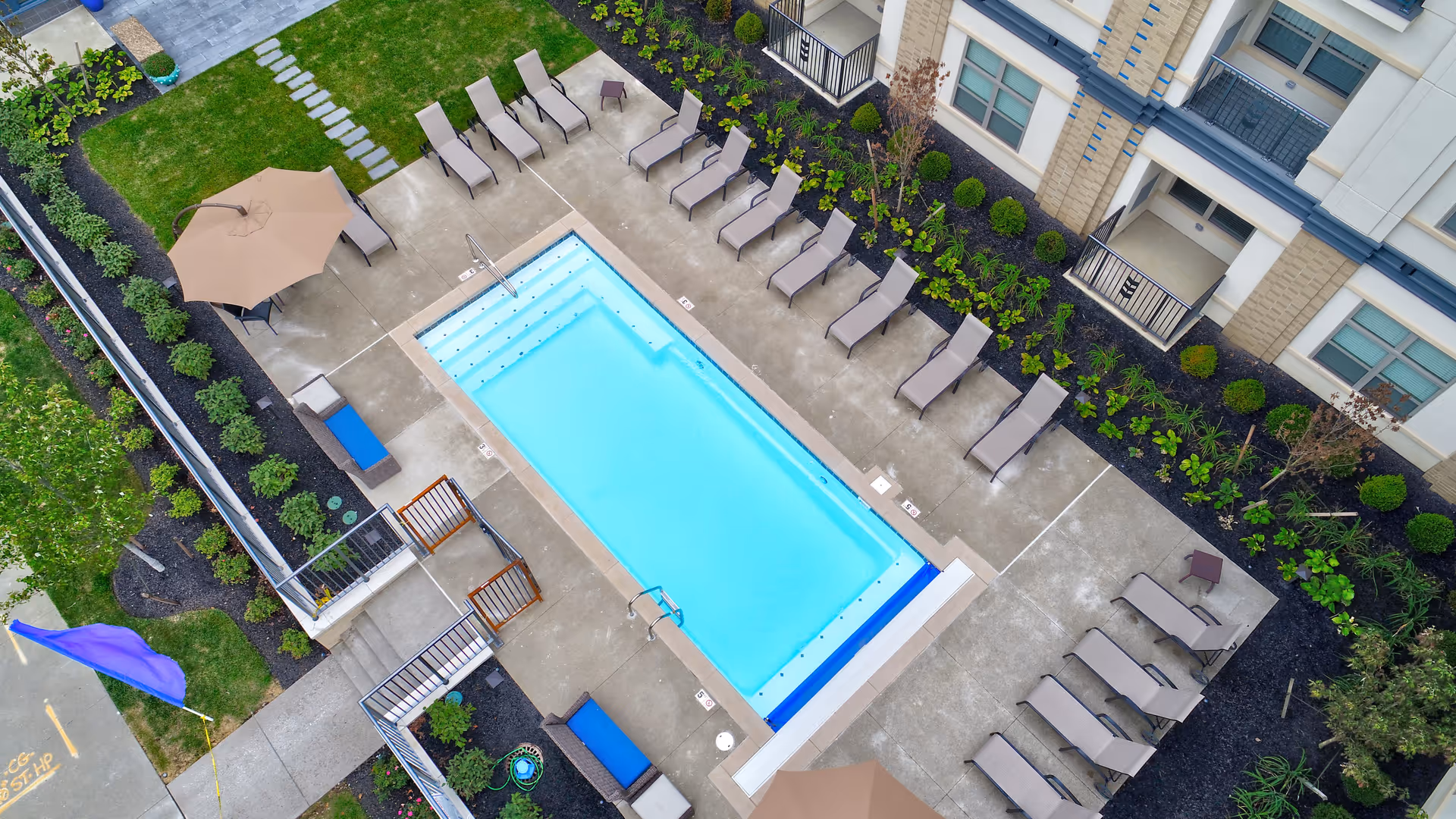 Aerial view of a rectangular swimming pool surrounded by lounge chairs, umbrellas, and landscaping in a courtyard next to a multi-story building.
