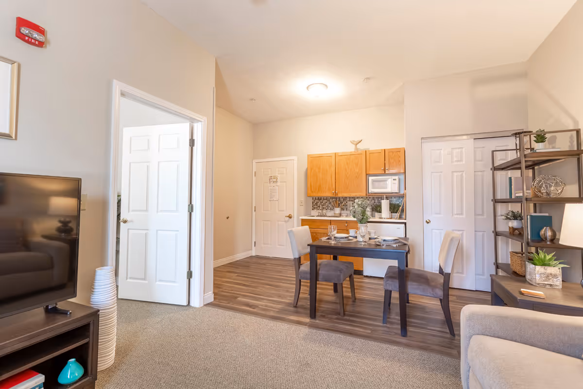 A bright and cozy living area in a senior living facility featuring a small dining table set for two, a kitchenette with wooden cabinets, a microwave, and a refrigerator. The room has a mix of carpet and wood flooring, a TV on a stand, a beige armchair, and a shelving unit with decorative items and plants. There are two white doors, one open and one closed, and a fire alarm on the wall.
