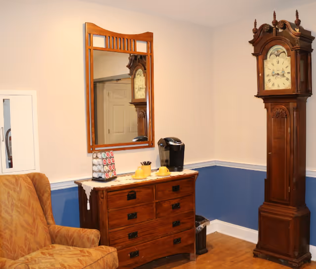 A cozy corner of a room featuring a wooden grandfather clock, a wooden dresser with a coffee maker and two yellow cups on top, a mirror hanging above the dresser, and a patterned upholstered armchair. The walls are painted beige on the upper half and blue on the lower half with white trim.