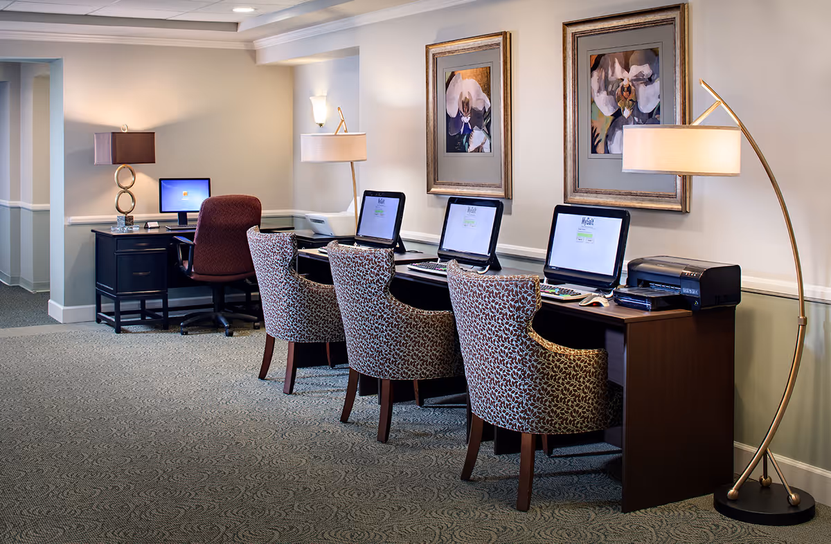 A senior living facility computer room with a row of three desktop computers on a long wooden desk, each paired with a patterned upholstered chair. There is a printer on the right side of the desk and two framed floral paintings on the wall above. To the left, there is another desk with a computer and a brown office chair. The room is softly lit with two floor lamps and a table lamp, and has carpeted flooring.