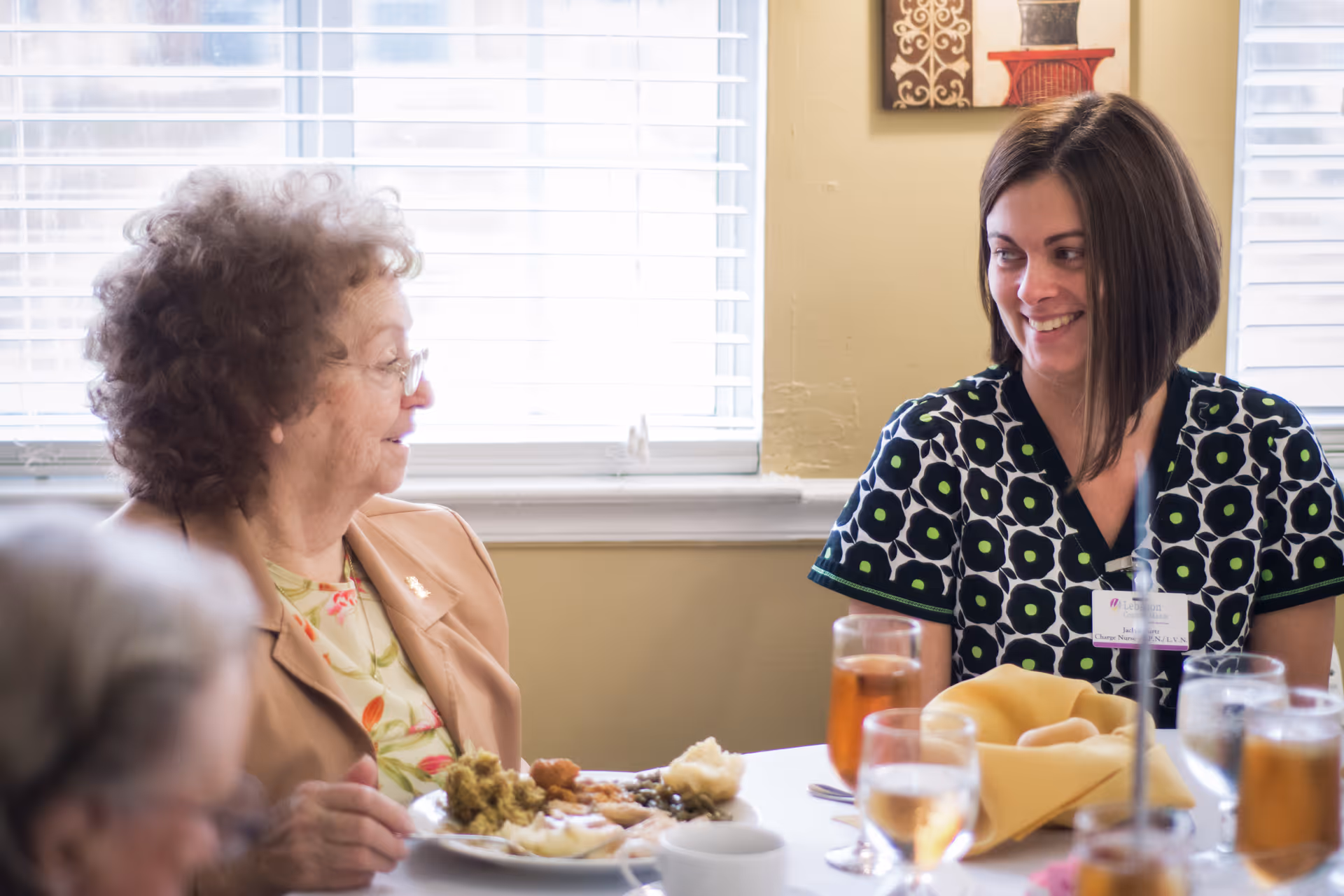 An older woman and a caregiver smile at each other while seated at a dining table with plates and glasses.