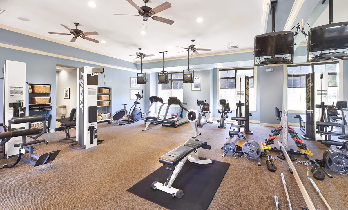 A well-lit fitness room with various exercise equipment including weight machines, stationary bikes, treadmills, free weights, and kettlebells. The room has large windows, ceiling fans, and mirrors on one wall.