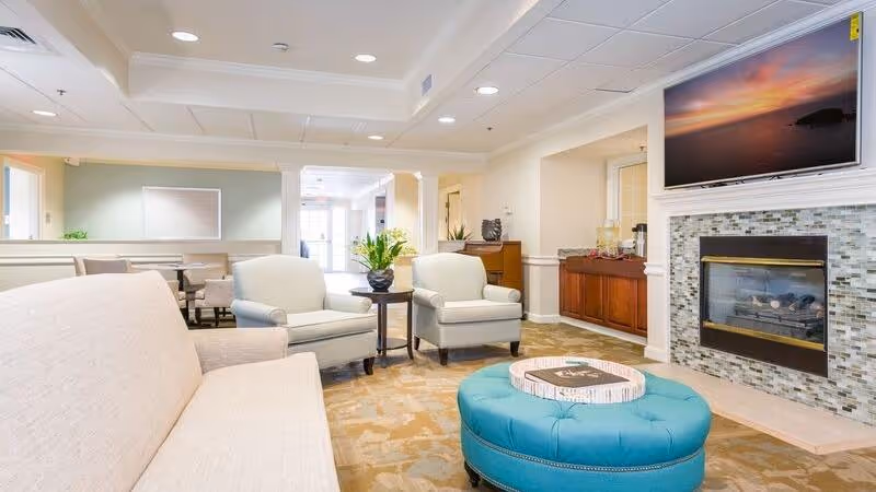 A bright and spacious living room area in a senior living facility featuring a beige sofa, two light-colored armchairs, a round teal ottoman with a tray on top, a fireplace with a tiled surround, and a large flat-screen TV mounted above the fireplace. There is a small table with a plant between the armchairs and a wooden cabinet with refreshments in the background.