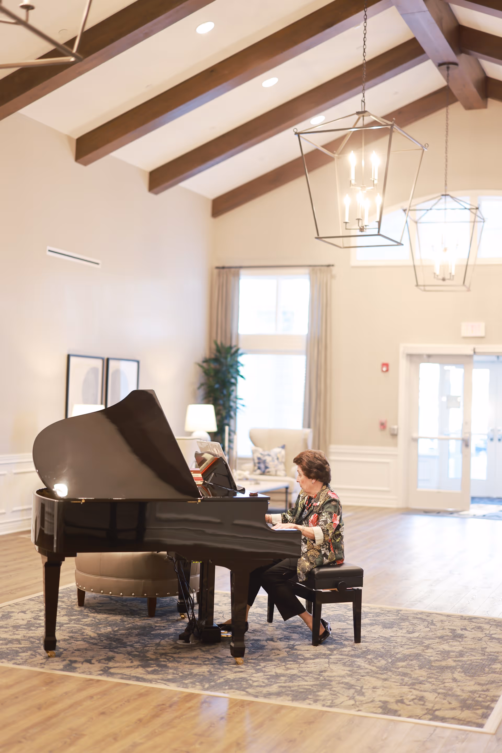An elderly woman playing a black grand piano in a spacious, well-lit room with wooden beams on the ceiling. The room features large windows with curtains, a patterned rug under the piano, and comfortable seating in the background.