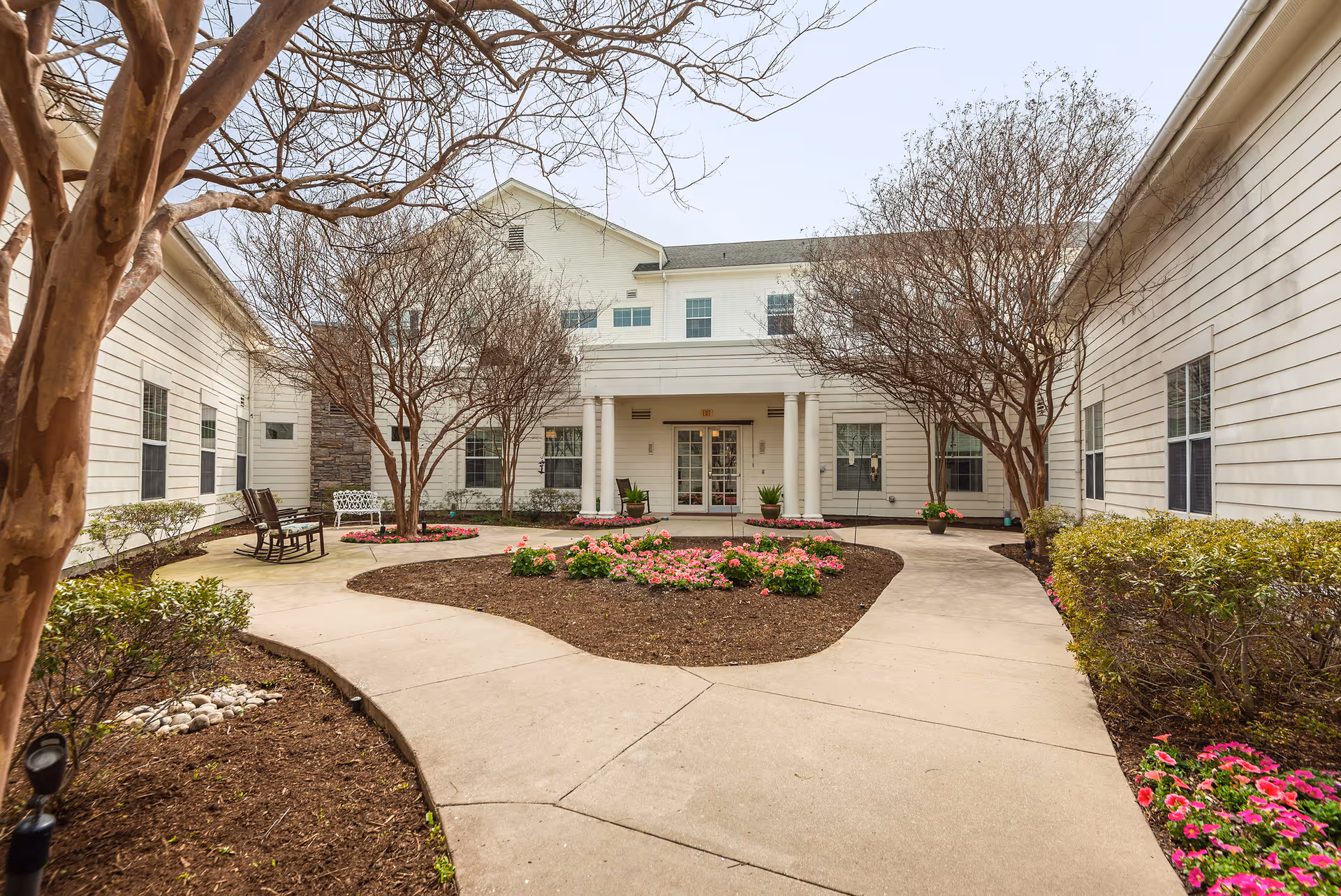 Outdoor courtyard area at Prestonwood Court featuring a paved walkway curving around landscaped flower beds with pink flowers and small trees. The courtyard is surrounded by a white two-story building with multiple windows and a central entrance with double glass doors and columns.