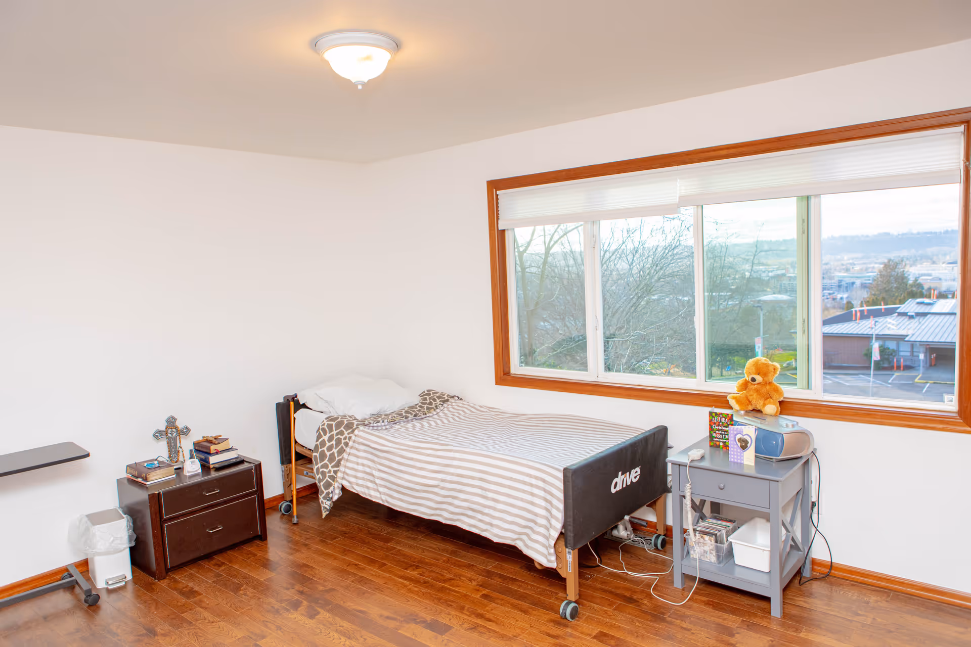 A bright single-occupancy bedroom with a striped bed, two bedside tables, a large window with views, and a teddy bear on a nightstand.