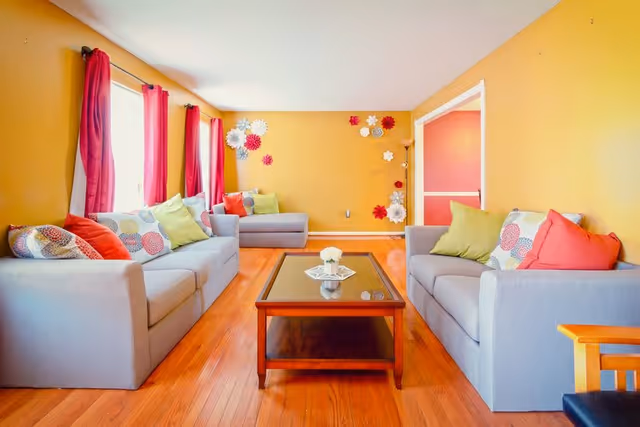 A bright living room with yellow walls, two gray sofas with colorful pillows, a wooden coffee table with a small flower arrangement, red curtains on the windows, and decorative wall flowers.