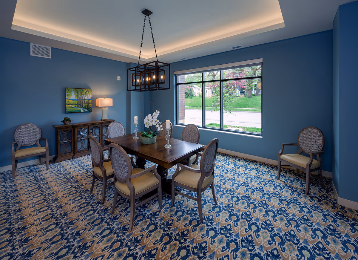 Blue-walled dining room with a wooden table and six chairs, a patterned carpet, large window, and a rectangular chandelier.
