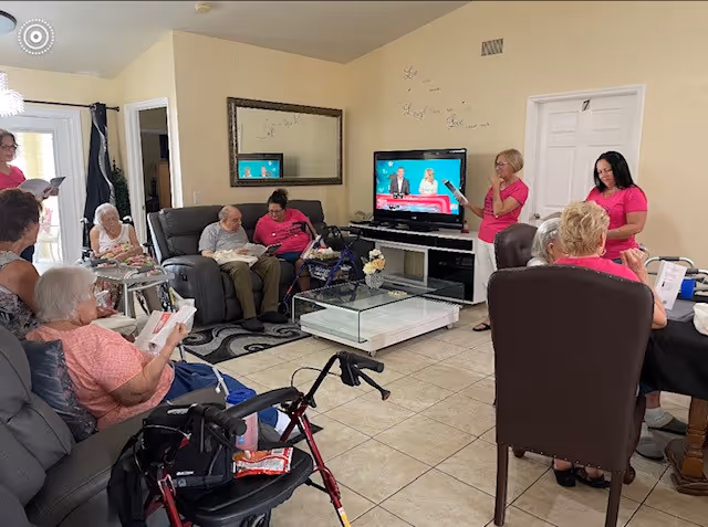 A group of elderly residents and staff gathered in a living room area of a senior living facility. Several residents are seated on sofas and chairs, some using walkers, while staff members in pink shirts stand and interact with them. A television is on, mounted on a stand against the wall, and a large mirror hangs above a sofa. The room has tiled floors and light-colored walls.