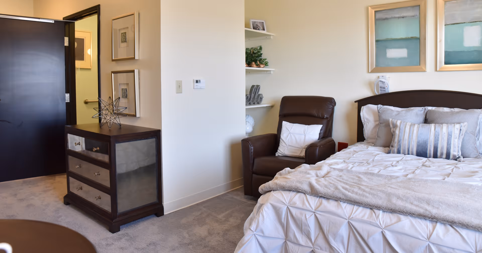 A cozy bedroom in a senior living facility featuring a neatly made bed with multiple pillows and a light-colored comforter. Next to the bed is a dark brown leather recliner chair with a white pillow. On the wall above the bed are two framed abstract paintings. A small dresser with drawers and decorative items is positioned near the entrance door, which is black. The room has light-colored walls and carpeted flooring.