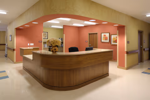 Reception desk area in a senior living facility with a wooden counter, a vase with dried flowers, and framed artwork on the walls. The walls are painted in warm tones of beige and terracotta, and the floor is light-colored with some blue square accents.