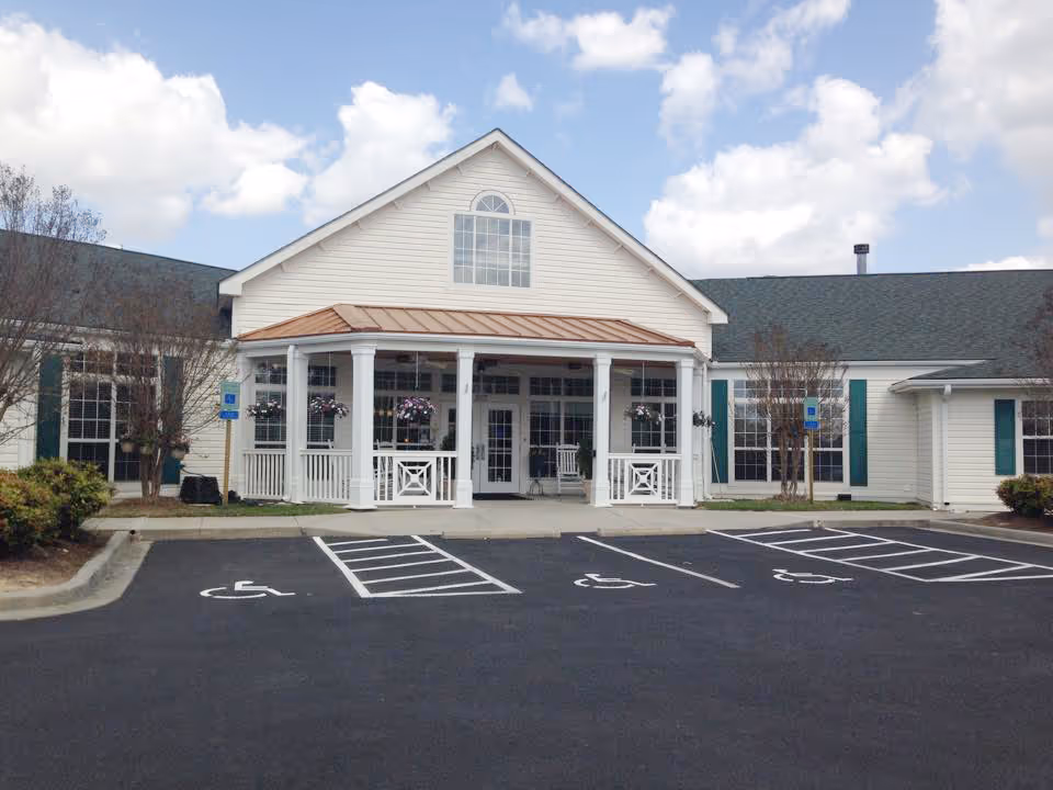 Front entrance of a single-story senior living facility with a covered porch, hanging planters, and marked handicap parking spaces in front.