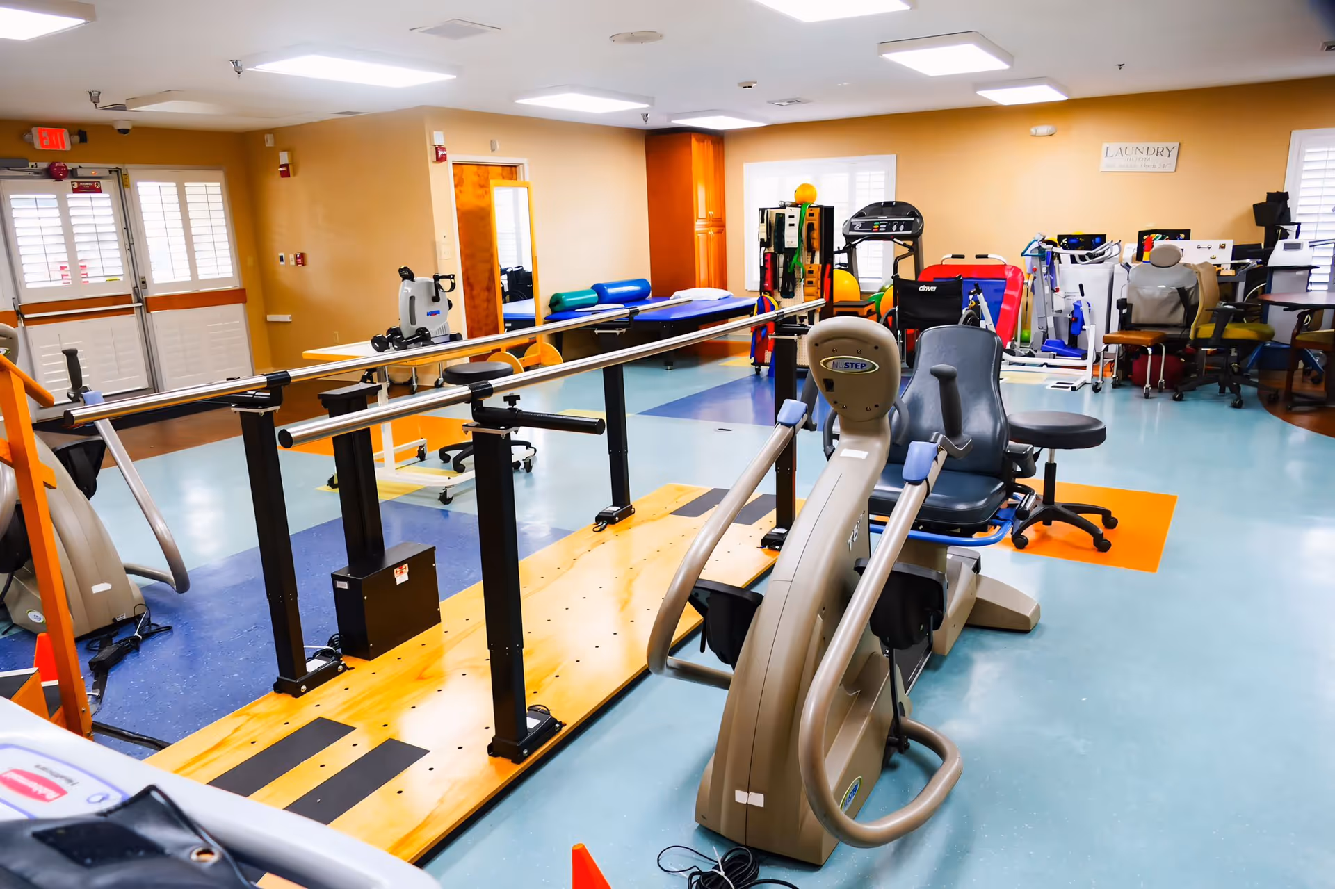 A rehabilitation center room equipped with various exercise and therapy equipment including parallel bars for walking practice, stationary exercise bikes, therapy chairs, and other physical therapy tools. The room has a light blue and orange floor, beige walls, and bright overhead lighting.