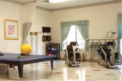 Therapy/exercise room with a treatment table, stationary recumbent bikes, exercise balls, and shelving under two curtained windows.