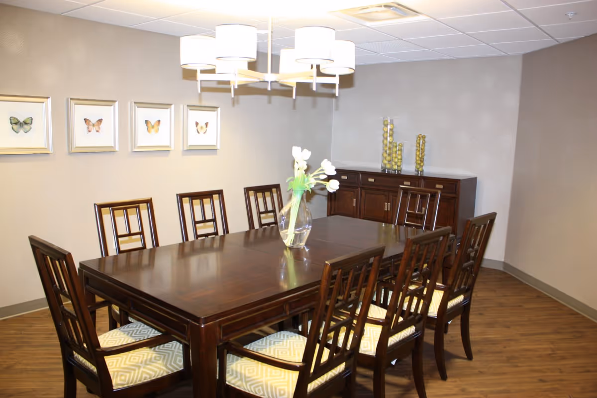 A dining room with a large dark wooden table surrounded by eight matching chairs with patterned cushions. A glass vase with white flowers is placed in the center of the table. On the wall behind the table, there are four framed butterfly pictures. A wooden sideboard with decorative glass containers filled with green balls is against the far wall. The room has wooden flooring and a modern ceiling light fixture with multiple lampshades.