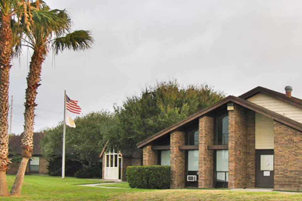 Exterior view of a brick building with large windows and a sloped roof, surrounded by green grass, palm trees, and other trees. Two flagpoles with the American flag and another flag are visible near the building.