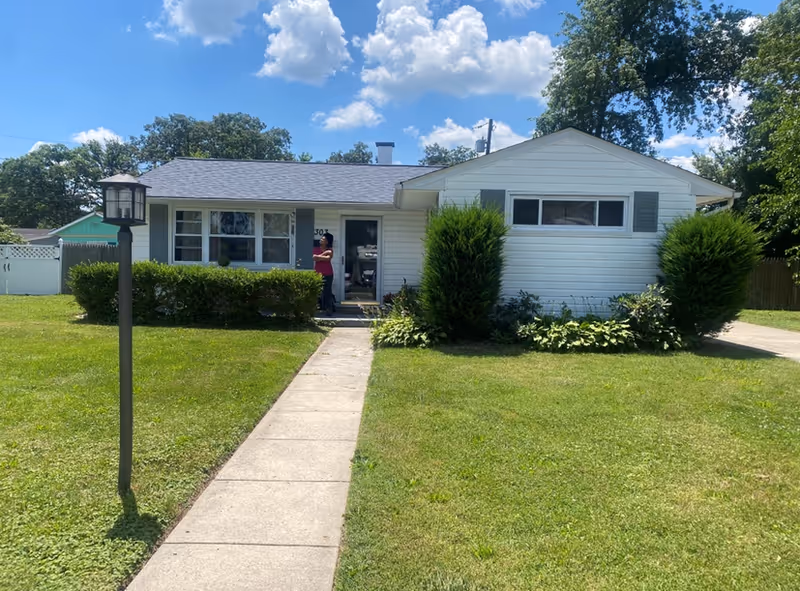 Single-story white house with a gray roof, a concrete walkway leading to the front door, green bushes and trees around the house, and a person standing near the entrance under a partly cloudy blue sky.