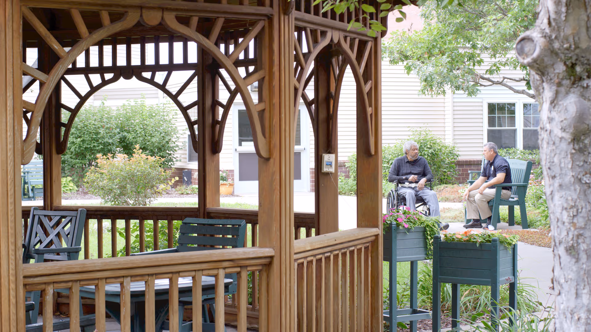 Two men sitting and talking outdoors near a wooden gazebo structure in a garden area with green plants and a building in the background. One man is in a wheelchair and the other is seated on a chair.