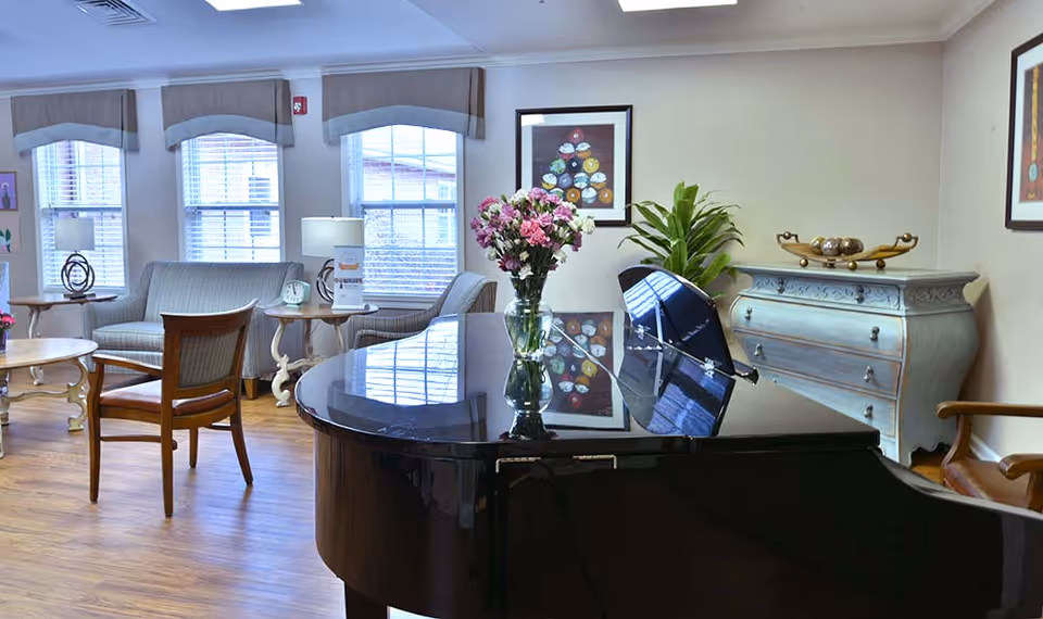 A bright and cozy living room area with a polished black grand piano in the foreground, topped with a vase of pink and white flowers. The room features wooden flooring, a light blue sofa, wooden chairs, side tables with lamps, and decorative framed artwork on the walls. A light blue chest of drawers with decorative items and a green plant are also visible.