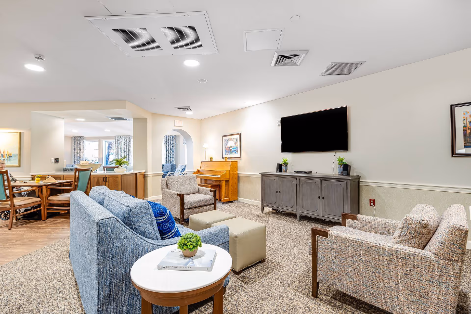 A bright and cozy living room area in a senior living facility featuring a blue sofa, two armchairs, a small round table with a plant and book, a piano against the wall, a large flat-screen TV mounted above a gray cabinet, and a dining area with wooden chairs and tables visible in the background.